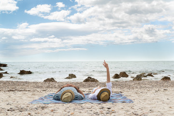 Young couple laying on a beautiful mediterranean beach and pointing at clouds. Relaxing time.