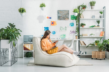 young woman using laptop and smartphone while sitting on soft chaise lounge in spacious room