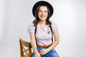 Young beautiful girl sitting on wooden chair in stylish black hat on white background. Emotions and gestures.