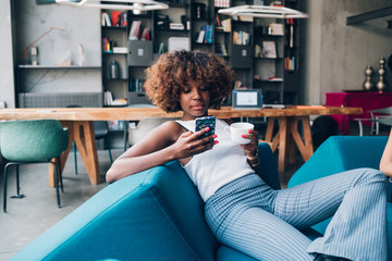 young black woman drinking and chatting with smartphone in modern loft