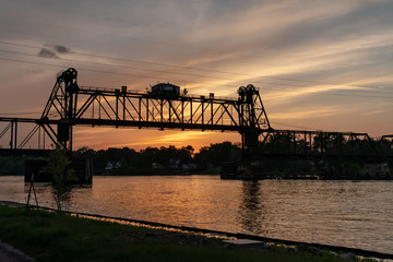 Obraz premium A movable, vertical-lift railroad trestle bridge over a river at sunset. Concepts of industry, transportation, urban landscape