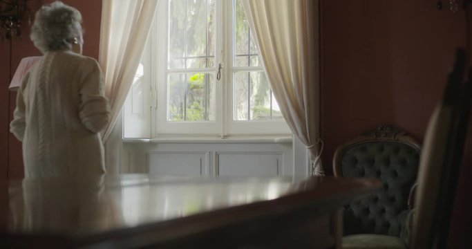 Senior grandma woman walking near window looking outside.Beautiful white hair elderly grandmother at home.4k slow motion video