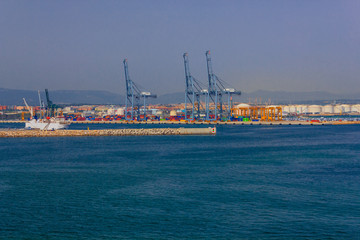 View of the commercial port of Tarragona in Spain with cranes to unload containers from merchant ships,  breakwater stones, and  the  warehouses for  storing goods  