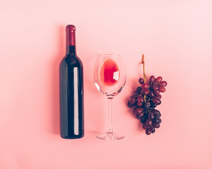 A bottle of wine an empty glass of a bunch of red grapes on a gentle pink background. Flat layout...