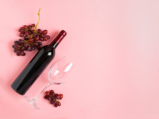 A bottle of wine an empty glass of a bunch of red grapes on a gentle pink background. Flat layout...