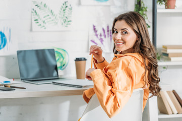 cheerful girl smiling at camera while sitting at desk near laptop with blank screen