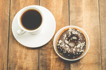 Сoffee break. White cup with black coffee and donat in chocolate glaze. Wooden background, top view.