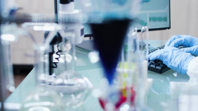 Dolly shot of modern high end laboratory elements, tubes and colored liquids. A scientis in glowes is typing on the computer keyboard