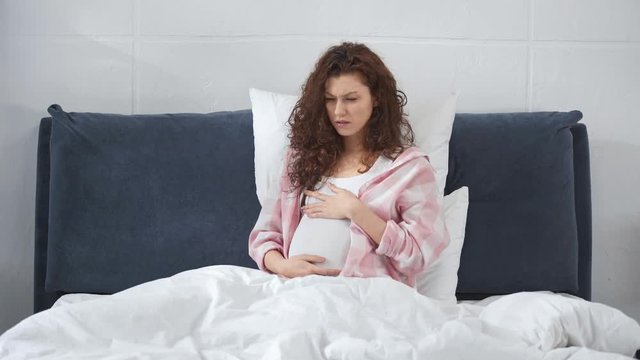pregnant young woman lying in bed, touching tummy and having toxicosis with nausea at home