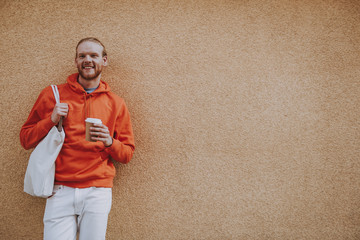 Happy red haired male leaning on wall with coffee