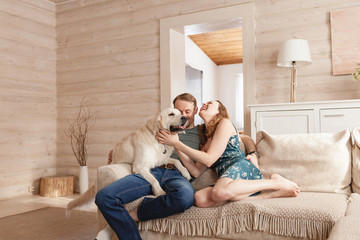 Young happy married couple nice girl and young man in casual clothes talking with his kind obedient dog in the living room of his cozy country house