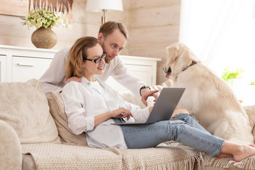 Happy newlyweds looking for a place for a summer holiday sitting on a sofa in their cozy living room next to their beloved dog