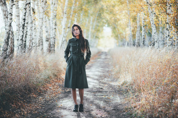 Sad beautiful girl with long natural black hair on bokeh background of autumn yellow leaves. Depressed girl in fall forest. Autumn apathy. Female emotional gloomy portrait in full growth. Faded tones.