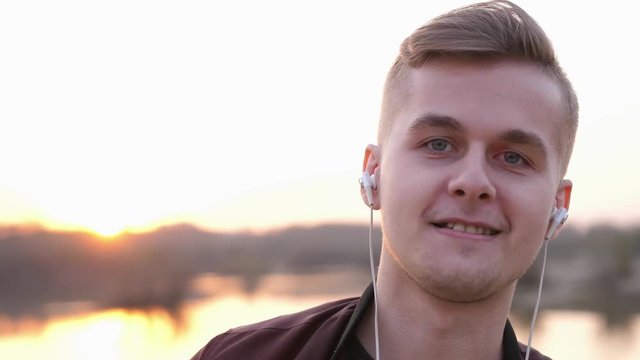 Young smiling handsome man listens to music looks at camera on beach during sunset. slow motion