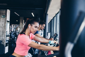 Young and attractive woman and man biking in the fitness gym. They exercising legs and doing cardio workout while riding cycling machines.