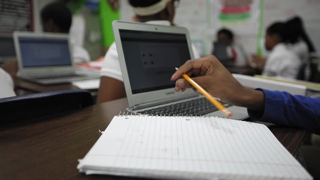 Close up of an African American high school student writing answers on paper with an open laptop computer.