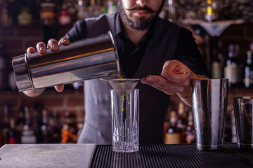 Bartender pouring a cocktail from the steel shaker on the bar counter on the blurred background