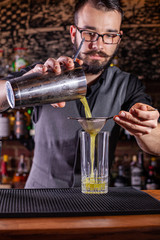 Bartender pouring a cocktail from the steel shaker on the bar counter on the blurred background