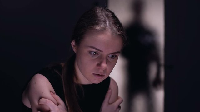 Scared crying woman sitting alone in room, cruel tyrant husband shadow outside