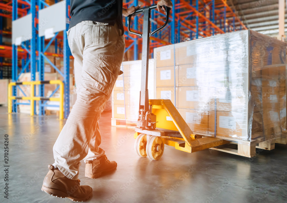 Wall mural warehouse worker unloading hand pallet jack with packaging boxes. supply chain in storage warehouse 