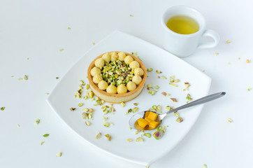 Modern tart with tropical flavor decorated with pistachio, spoon with mango pieces and cup of green tea on white plate on white background