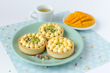 Modern tarts with tropical flavor decorated with pistachio, cup of green tea and fresh mango on green plate on white background