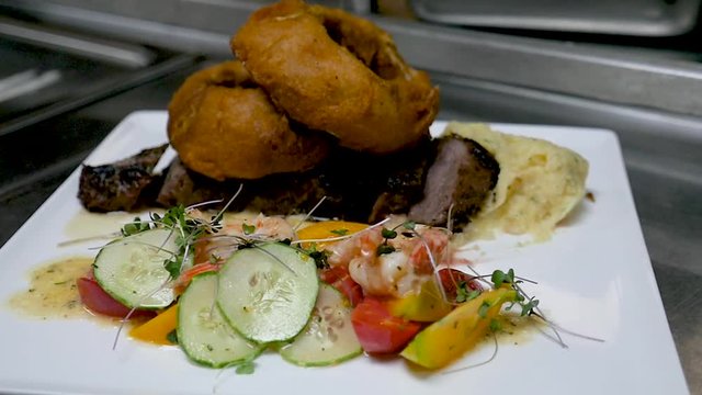 chefs special steak and oinion rings in kitchen. The camera turns around a chef special dish in a stainless steel kitchen. The dish is steak with large onion rings on top and veggies on the side.
