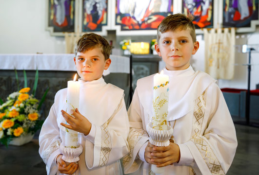 Two little kids boys receiving his first holy communion. Happy children holding Christening candle. Tradition in catholic curch. Kids in a church near altar. Siblings, brothers in white gowns.