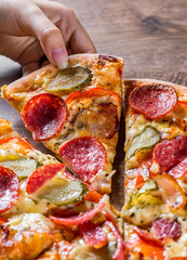 female hand takes a slice of Pepperoni Pizza with Mozzarella cheese, salami, bacon, Tomato sauce, pepper, Spices and pickled cucumbers. Italian pizza on wooden table background