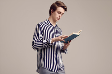 Young handsome man in striped shirt reading a book on gray background