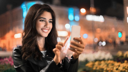 Happy woman sending message on phone, walking in night city
