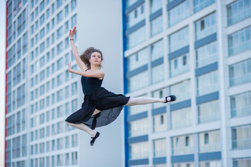 Ballerina in a tutu posing at a multi-storey residential building. Beautiful young woman in black dress and pointe shoes outside. Gorgeous ballerina performs elegant jumps.