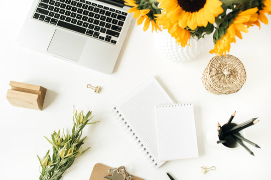 Home office desk workspace with laptop, notebook, clipboard, yellow sunflowers bouquet on white background. Flat lay, top view freelancer / blogger work concept.