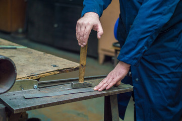 Professional locksmith in blue uniform working with hammer and steel at crafting table - metalworking industry concept. 