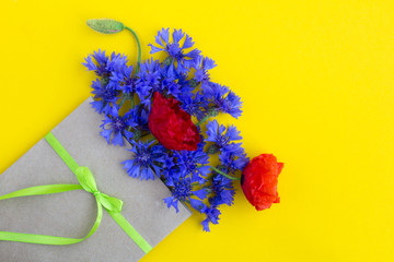 Bouquet of red poppies and blue cornflowers in a envelope tied with a green ribbon on the yellow background.Top view.Copy space.Spring or summer flowers concept.