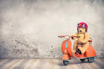 Retro Teddy Bear toy in red helmet with goggles sitting on old children's pedal orange scooter from 60s front loft concrete wall background. Kid racer concept. Vintage style filtered photo