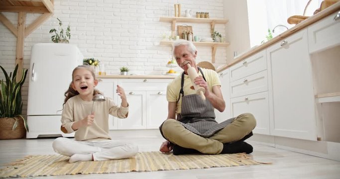 Little Girl Playing With Her Caucasian Granddad In Kitchen, Singing Into A Ladle And Smiling - Grandfather Supporting Childhood Dreamof Kid - Family Ties, Family Time Concept 4k