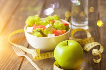 Salad in white bowl, fresh green apple and tape measure on dark wooden background. Diet and weight loss concept.