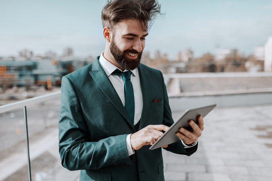 Young smiling successful bearded businessman in formal wear standing on the rooftop on pause and using tablet.