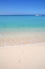 Footprints leading out of the ocean on a sandy tropical beach with a boat in the background