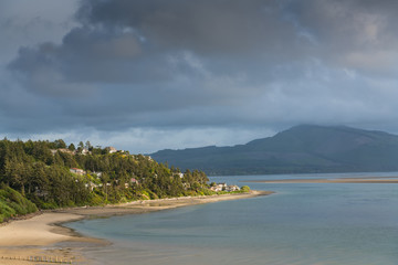Fototapeta premium Light from sunset spotlights homes and a golden sandy beach along a calm bay under dramatic clouds at Netarts, Oregon