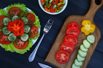 A cutting board with slices of tomatoes, cucumbers and pepper, a fork and a knife and two plates of vegetable salads, healthy food, dark wooden background, diagonal, top view, closeup