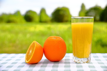 A glass of orange juice with oranges on a green-and-white checkered tablecloth, blurred green natural background on a sunny day