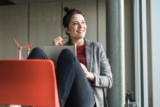 Smiling businesswoman sitting on chair in office with laptop looking sideways