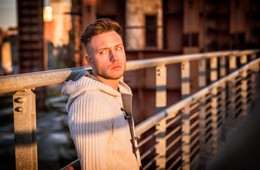 Handsome trendy young man, standing on a sidewalk in city setting at sunset wearing a fashionable sweater or cardigan