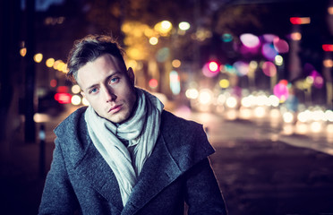 Handsome trendy young man, standing on a sidewalk in city setting at night wearing a fashionable winter coat and scarf