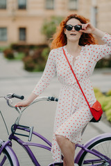 Street style and active rest concept. Curly red haired woman keeps hand on frame of sunglasses,...