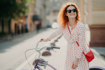 Smiling red haired European woman has glad expression, stands near bicycle, carries red bag,...