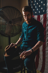 A young man with full beard and felt hat sitting in front of an America flag in a distressed room.