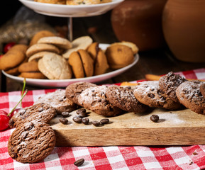 Oatmeal chocolate cookies with coffee grains and cherry, powdered sugar on kitchen cutting board with checkered fabric on wooden table in village style for picnic. Useful for digestion.